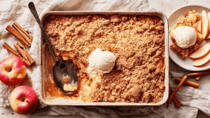 Apple Crumble kalorienarm im weißen Backblech mit goldbraunen Streuseln und Vanilleeis, schwarzer Servierlöffel im Crumble, frische rote Äpfel und Zimtstangen als Deko, Teller mit serviertem Dessert rechts, rustikale Leinendecke
