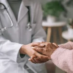 Ärztin hält empathisch Hand einer Patientin bei Brustkrebsvorsorge-Beratung - vertrauensvolle Arzt-Patienten-Beziehung im medizinischen Gespräch