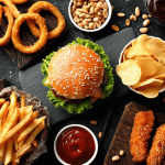 Verschiedene Fast-Food-Gerichte von oben fotografiert: Pizza, Burger, Pommes, Zwiebelringe und Snacks auf dunklem Schiefer