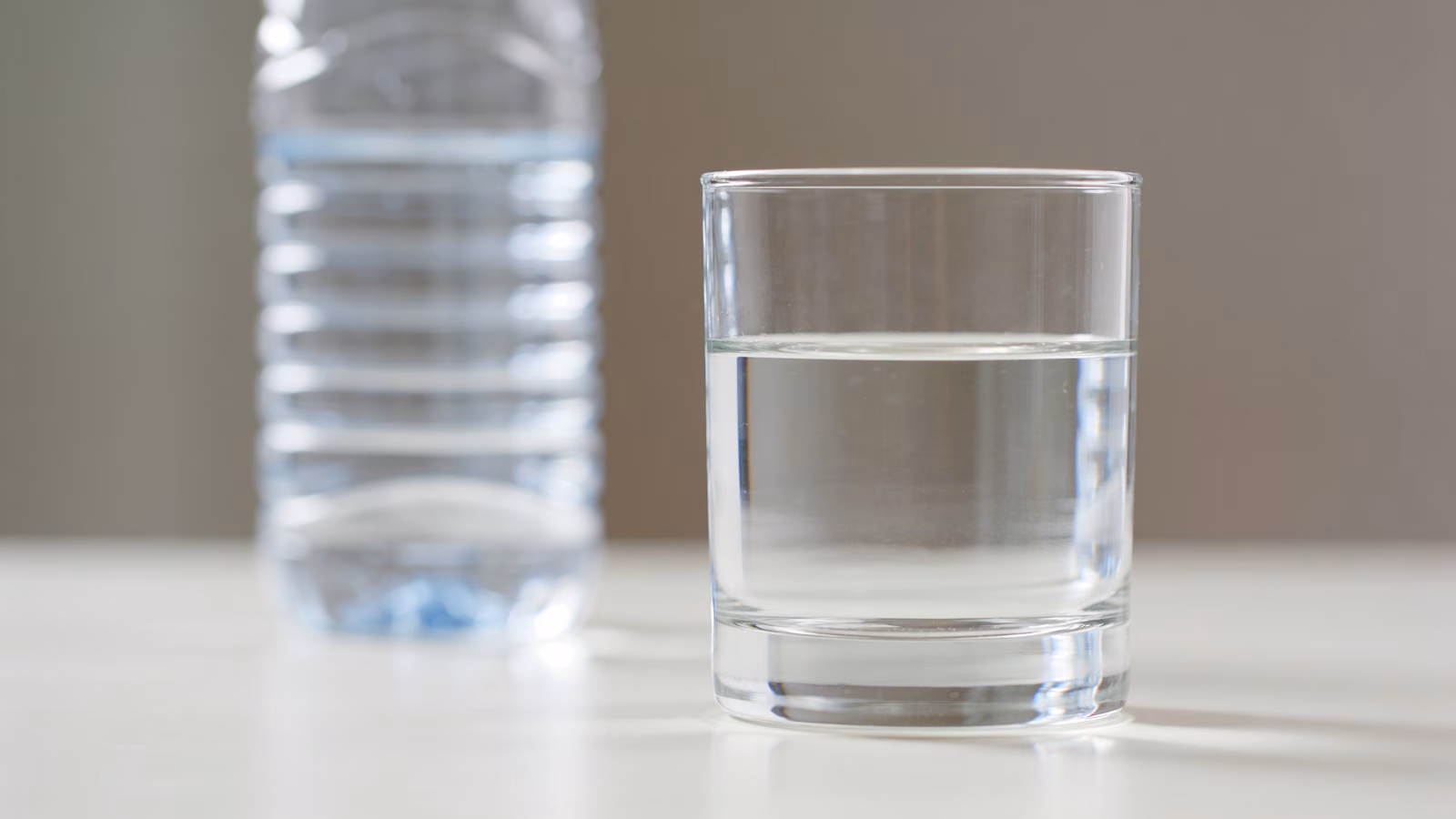 Ein Glas Wasser vor einer unscharfen Plastikflasche auf hellem Untergrund